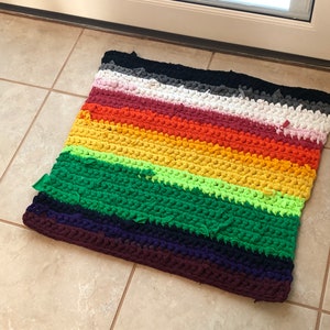 May include: A multi-colored striped crocheted doormat with a rainbow color scheme. The doormat is made of thick yarn and has a textured surface.