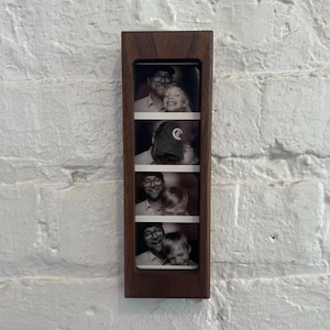 May include: A dark wood photo frame with four black and white photo booth style images. The images show a man and a child in various poses. The frame is rectangular and mounted on a white brick wall.