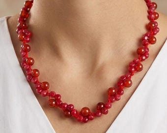 May include: A necklace featuring a series of small, vibrant red beads intertwined with larger, translucent red spherical beads. The beads are strung together in a clustered, textured pattern that sits around the neck, creating a continuous, circular design.