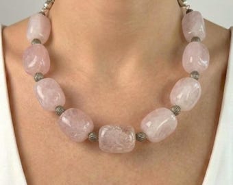 May include: A necklace featuring large, polished rose quartz beads in varying sizes. The light pink beads are separated by small, silver-colored metal beads. The necklace is worn around the neck.