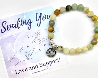 May include: A green, brown, and cream beaded bracelet with a silver charm featuring a lotus flower. The bracelet is on a white background with a card that says "Sending You Love and Support!"