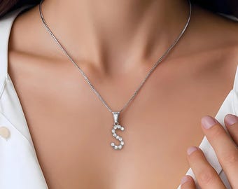 May include: A silver-toned necklace featuring a pendant shaped like the letter S, encrusted with small, clear, sparkling stones. The pendant hangs from a delicate, thin silver-toned chain, resting against a plain white collared shirt.