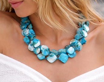 May include: A statement necklace featuring blue and white shell-like beads. The necklace is worn with a white, off-the-shoulder cardigan with silver buttons. The cardigan is paired with blue jeans.