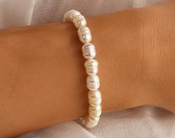 May include: A white pearl bracelet. The bracelet is made of a series of white pearls strung together. The bracelet is worn on a person's wrist. The background is a white shirt.