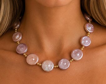May include: A necklace featuring a series of round, iridescent lavender coin-shaped pearls alternating with small, polished gold-toned spherical beads. The pearls exhibit a smooth, lustrous surface with subtle variations in light purple tones.