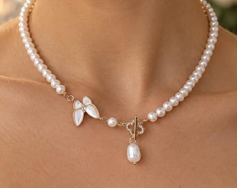 May include: A white pearl necklace featuring a small white butterfly charm and a gold-toned toggle clasp with a single dangling baroque pearl. The necklace is composed of uniform round white pearls and includes a small clover-shaped metal accent.