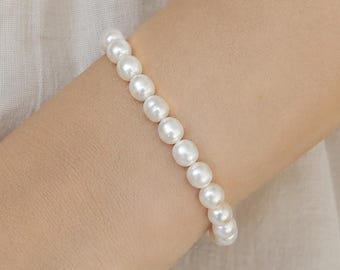 May include: A white pearl bracelet. The bracelet is composed of round, lustrous white pearls. The pearls are strung together, forming a delicate and elegant piece of jewelry. The bracelet is shown on a person's wrist.