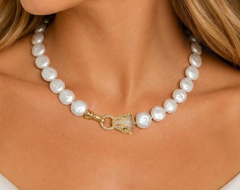 May include: A necklace featuring a single strand of round white pearls. The necklace is secured with a gold-toned clasp shaped like a stylized animal head, which is encrusted with small, clear, sparkling stones at the center front.
