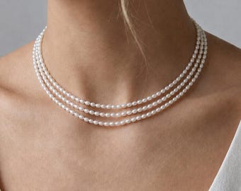 May include: A triple-strand pearl necklace. The necklace features three delicate strands of small, round, white pearls. The necklace is worn against a white top, creating a classic and elegant look. The pearls are evenly spaced and have a subtle luster.