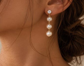 May include: A gold drop earring featuring a single clear round stud at the top, followed by a vertical arrangement of three white spherical pearls suspended on a thin gold chain. The earring is secured to the earlobe with a gold post backing.