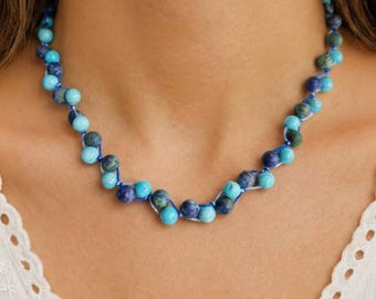 May include: A beaded necklace featuring a series of round, polished stones in varying shades of turquoise, sky blue, and deep navy blue. The stones are strung together on a thin, vibrant blue cord, creating a textured, multi-colored circular design.