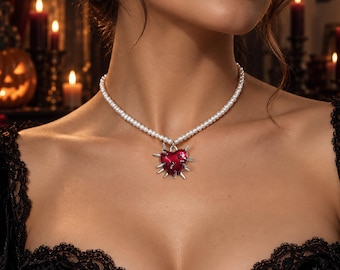 May include: A necklace featuring a strand of small white pearls and a central pendant shaped like a red heart. The heart pendant is accented with multiple silver-toned spikes protruding from its edges, creating a distinct contrast in texture.