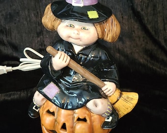 May include: A ceramic figurine of a witch riding a pumpkin. The witch is wearing a black hat with a purple band, a black dress, and black shoes. She is holding a brown broom in her hands. The pumpkin is orange and has a carved face.