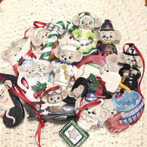 May include: A collection of ceramic mouse ornaments, each with unique designs and costumes. The ornaments include a chef, pilot, and firefighter, with red ribbon hangers. The ornaments are on a cream-colored, crocheted surface.