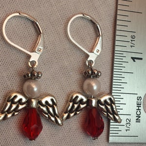 May include: Silver dangle earrings with a red crystal and a white pearl, shaped like an angel with wings.