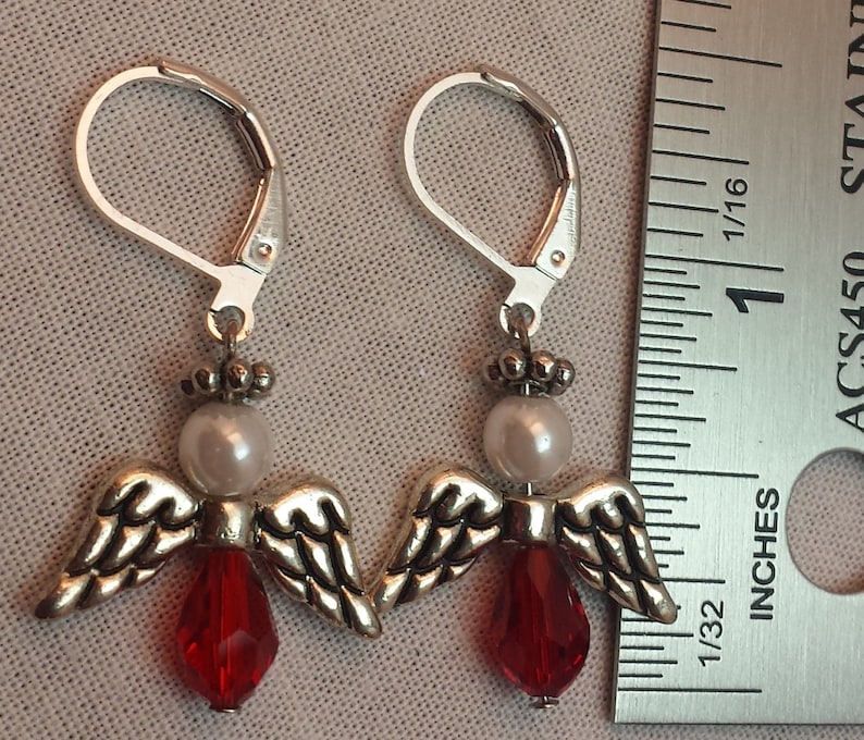 May include: Silver dangle earrings with a red crystal and a white pearl, shaped like an angel with wings.