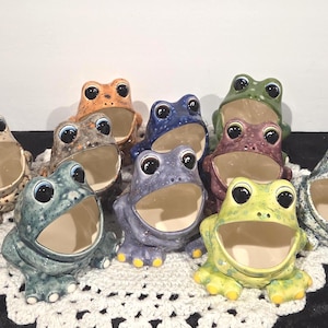 May include: A collection of ten ceramic frog-shaped containers, each featuring a wide open mouth, large black eyes, and a unique speckled or mottled glaze in various colors including green, blue, purple, orange, brown, and cream.