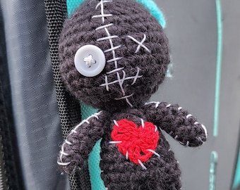 May include: A handmade black crochet doll keychain with white stitching and a red heart. The doll has a button eye and a small tuft of blue yarn. The keychain is attached to a silver ring.