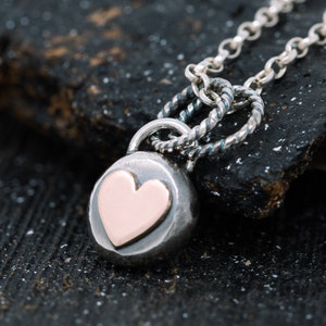 May include: A silver necklace with a round pendant featuring a rose gold heart. The necklace has a twisted chain and a lobster clasp.