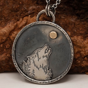 May include: A silver pendant necklace featuring a howling wolf silhouette against a dark gray background with a gold-colored moon. The pendant is round with a textured border and hangs from a silver chain. The design includes a forest scene.
