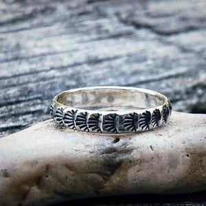 Könnte beinhalten: Ein silberner Ring mit einem sich wiederholenden Muster aus stilisierten fächerartigen Formen. Der Ring liegt auf einem hellgrauen Stein, mit einem verschwommenen Holzgrund. Das Design des Rings ist detailliert und das Metall hat eine polierte Oberfläche.