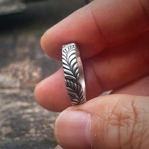 May include: A silver ring with a leaf pattern. The ring is held between fingers, showcasing the detailed design. The ring's surface is etched with a repeating pattern of stylized leaves, creating a textured appearance. The ring is a piece of jewellery.