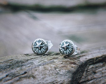 May include: A pair of silver star stud earrings. The earrings are round and have a star design in the center. The earrings are on a wooden surface.