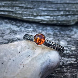 May include: A silver ring featuring an amber gemstone. The ring's band is intricately engraved with a decorative pattern. The amber stone is a warm, orange-brown hue and is set in a simple bezel. The ring is displayed on a light-colored stone.