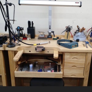 May include: A wooden workbench with a variety of tools and supplies for jewellery making. The workbench has a drawer with tools and a magnifying head lamp. There is a lamp with a long arm and a fluorescent light.