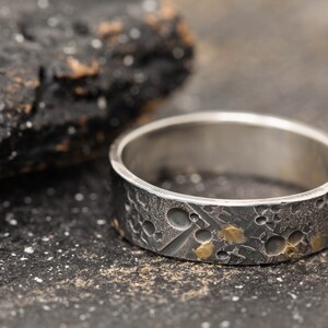 May include: A silver ring with a textured surface and gold accents. The ring has a cratered design, resembling the surface of the moon.
