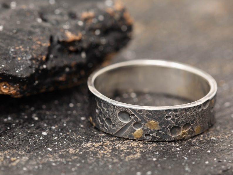 May include: A silver ring with a textured surface and gold accents. The ring has a cratered design, resembling the surface of the moon.