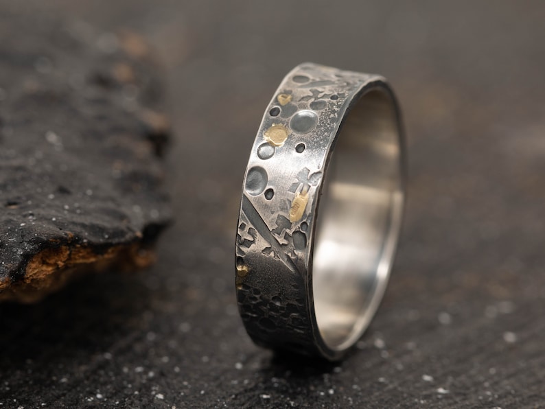 May include: A silver ring with a textured surface and gold accents. The ring has a hammered finish and a unique design.