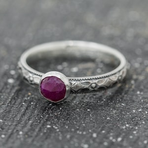 Könnte beinhalten: Ein silberner Ring mit einem verzierten Blumenmuster und einem tief roten Rubin-Edelstein.