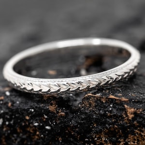 May include: A silver wedding band with a delicate, textured design. The band is thin and has a subtle, intricate pattern.
