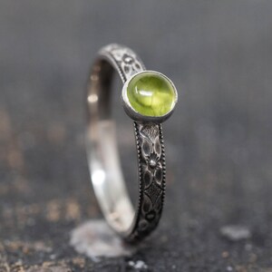 Könnte beinhalten: Ein silberner Ring mit einem runden, hellgrünen Edelstein. Das Band weist ein geätztes Blumenmuster auf. Der Ring ist vor einem dunklen, strukturierten Hintergrund platziert, der die Farbe des Edelsteins und das aufwendige Design des Rings hervorhebt.