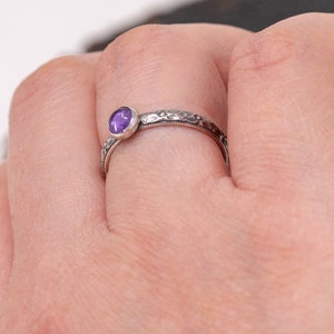 May include: A silver ring with a purple gemstone set in a bezel setting. The ring has a delicate, textured band.