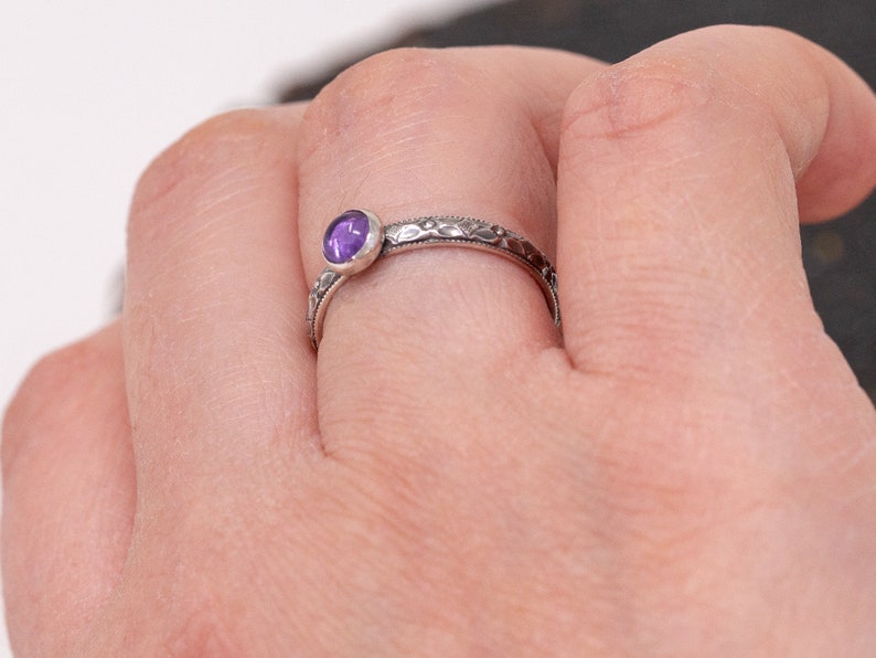 May include: A silver ring with a purple gemstone set in a bezel setting. The ring has a delicate, textured band.