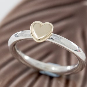 May include: A silver ring with a gold heart-shaped accent. The ring is smooth and shiny.