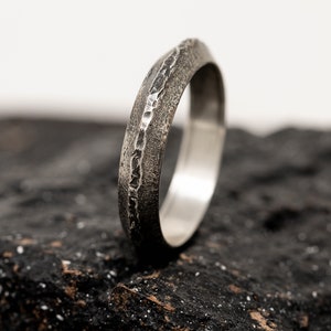 May include: A silver ring with a textured band and a smooth inner band. The ring is sitting on a dark grey surface.