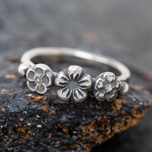 May include: A silver ring with three flower motifs. The ring has a smooth band, and the flowers are grouped together, forming a floral pattern. The ring is displayed against a dark, textured backdrop.