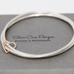 May include: A silver bangle bracelet with a gold heart charm. The bracelet is double-layered and has a polished finish. The charm is a simple heart shape and is attached to the bracelet with a small loop.