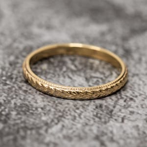 May include: A gold ring with a detailed, textured pattern. The ring has a repeating leaf-like design around the band, with a beaded border. The ring is likely made of gold and is a piece of jewellery.