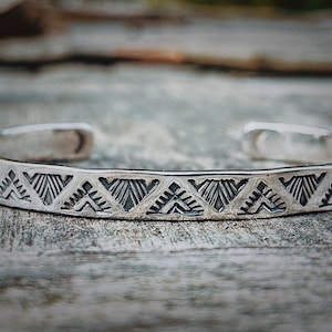 May include: A silver bracelet with a geometric pattern of triangles. The bracelet is made of a thin band of metal and has a simple, elegant design.