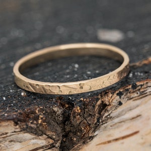 May include: A gold-colored ring with a textured surface, resting on a dark, textured surface. The ring has a hammered design, giving it a unique, handcrafted appearance. The ring is a simple band, perfect for everyday wear.