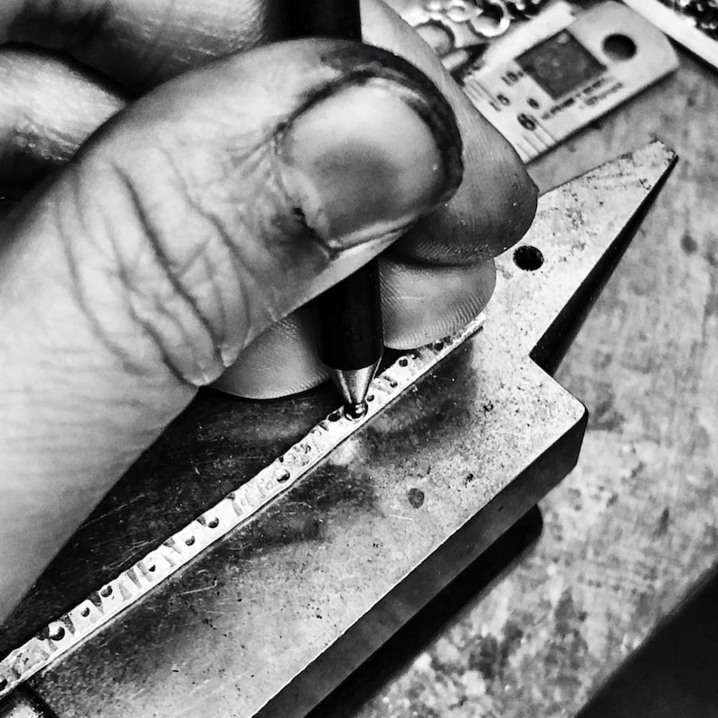 May include: A close-up of a hand using a tool to mark a metal surface. The surface has a series of small indentations.