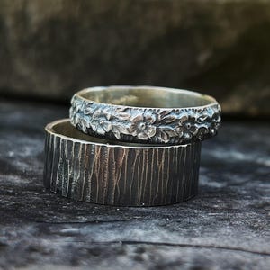 May include: Two silver rings. The top ring has a floral pattern around the band. The bottom ring has a textured, bark-like appearance. The rings are stacked, highlighting their contrasting textures and designs. The rings are made of silver.