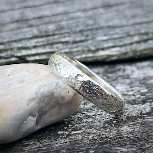 May include: A silver ring with a textured surface rests on a white stone against a weathered wood background. The ring's surface has a hammered appearance, reflecting light. The ring is a simple band design, suitable for everyday wear.