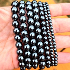 May include: A set of seven black hematite bead bracelets, each with a different size bead. The bracelets are stacked on top of each other, showing the different sizes.