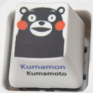 May include: A white keyboard key with a black and white cartoon bear with red cheeks. The text "Kumamon Kumamoto" is printed below the bear.