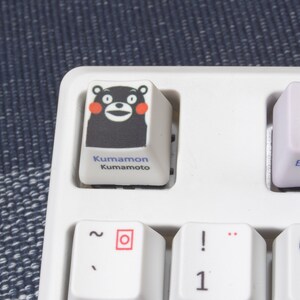 May include: A white keyboard with a black and white keycap featuring a cartoon bear with red cheeks. The keycap says "Kumamon Kumamoto".